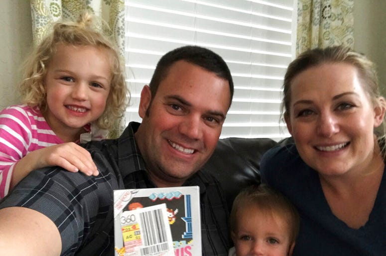 In this June 15, 2019 photo provided by Heritage Auctions, Scott Amos, along with his wife, Kristy, and daughters Grace, left, and Katie, pose in Reno, Nev., with an unopened copy of a 1987 cult-classic video game "Kid Icarus." The boxed game cartridge, s