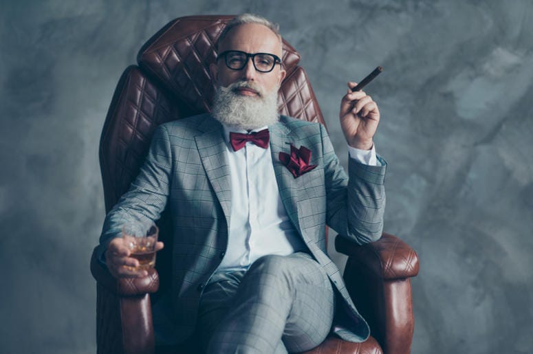 Cool man in glasses, hold cigarette, glass with brandy, in formal wear, tux with red bowtie and pocket square, sit in leather chair over gray background, looking to the camera, shares, stock, money