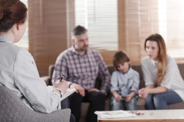 Rear view of female psychologist helping young family with a kid to solve child development problems. Family sitting on a sofa in the blurred background -
