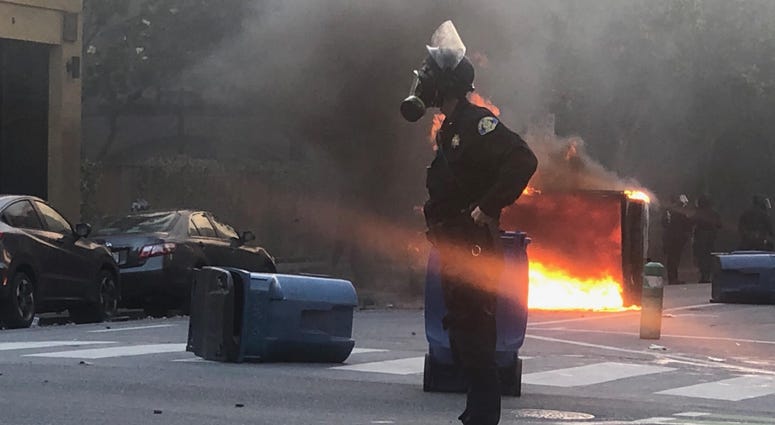 An officer stands alone as a dumpster burns in the background.