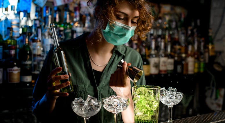 Young bartender girl in a medical mask pour green liquid from beaker into glass with ice. Young bartender girl in a medical mask pour green liquid from beaker into glass with ice.