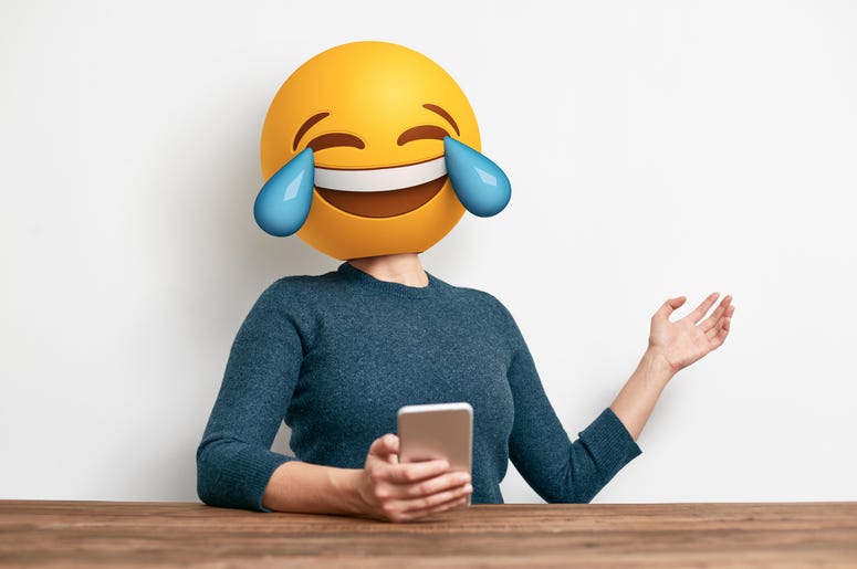 (atomicstudio/ iStock/ Getty Images Plus) Emoji Head Woman sitting at desk. Woman wearing tears of joy emoji masks while looking at her phone. This emoji is laughing so much that it is crying tears of joy