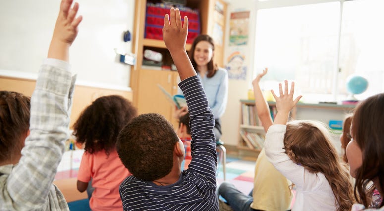 Elementary school children raising their hands in front of their teacher.