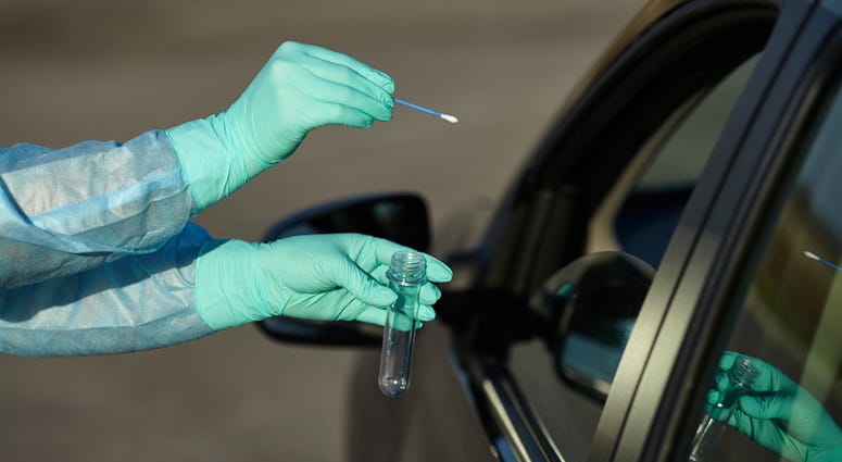Medical worker in full protective gear takes sample from patient at a COVID-19 drive-thru test site. Testing is done by throat swab.