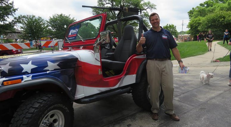 Kris Kobach stands smiling next to his patriotic-styled Jeep that has a replica machine gun mounted to it. He's giving the camera a thumbs up.