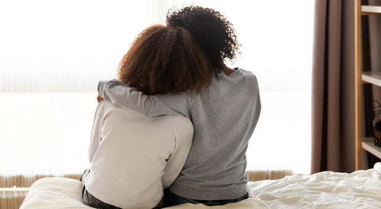 Two little girls, sitting on a bed, shown from behind, hugging each other.