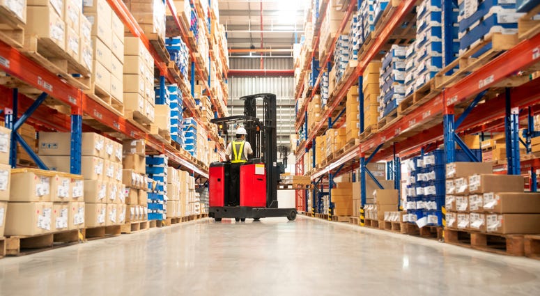 Worker in forklift-truck loading packed goods in huge distribution warehouse with high shelves.