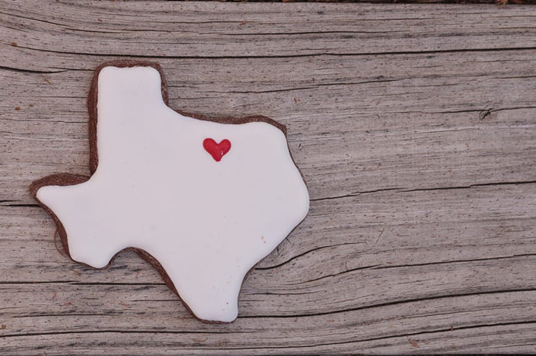 Texas Es Uno De Los Mejores Estado Para Solteros Texas shaped sugar cookie with white icing and a red heart on a rustic wood table.