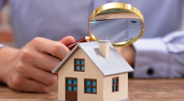 Close-up Of A Businessman's Hand Holding Magnifying Glass Over House Model Over Desk