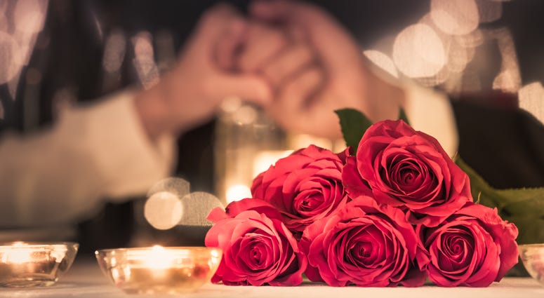 Couple at a candle light dinner date holding hands next to bouquet of red roses.