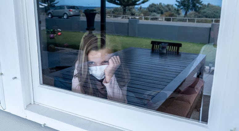 Depressed lonely little girl with face mask looking through the window during quarantine.