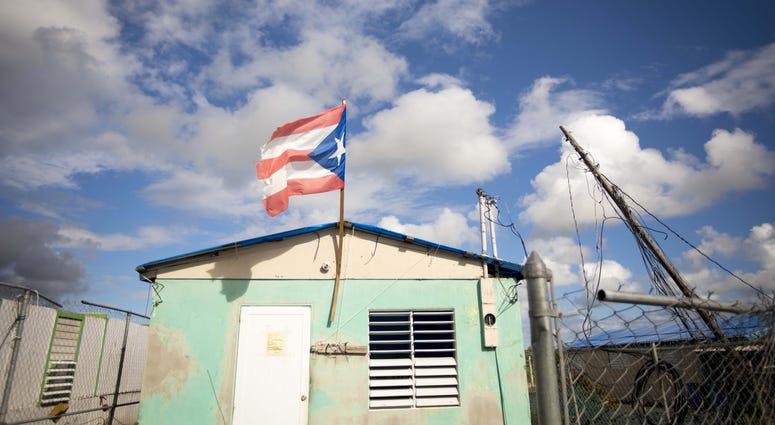 A house with a Puerto Rican flag and a blue tarp over the roof stands in Yabucoa, Puerto Rico, as an electrical post leaning behind it. Feb. 28, 2018. A house with a Puerto Rican flag Puerto Rico.