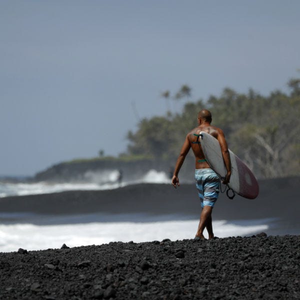 In this Tuesday, April 23, 2019 photo, a surfer walks on a newly formed black sand beach below Kilauea volcano near Kapoho, Hawaii In this Tuesday, April 23, 2019 photo, a surfer walks on a newly formed black sand beach below Kilauea volcano near Kapoho, Hawaii