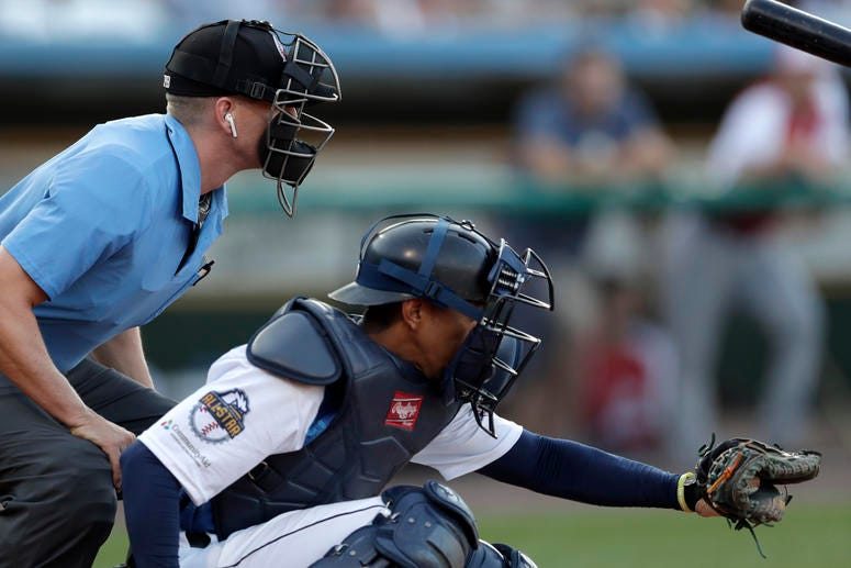 Home plate umpire Brian deBrauwere, left, huddles behind Freedom Division catcher James Skelton, of the York Revolution, as the official wears an earpiece during the first inning of the Atlantic League All-Star minor league baseball game, July 10, 2019. Umpire