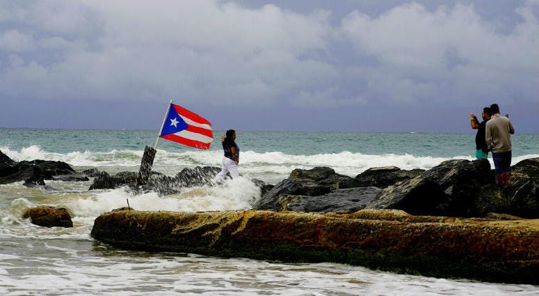 A woman poses for a photo backdropped by ocean waters and a Puerto Rican national flag, after the passing of Tropical Storm Dorian, in the Condado district of San Juan, Puerto Rico, Wednesday, Aug. 28, 2019. The Hurricane Center said the storm could grow into a dangerous Category 3 storm as it pushes northwest in the general direction of Florida.