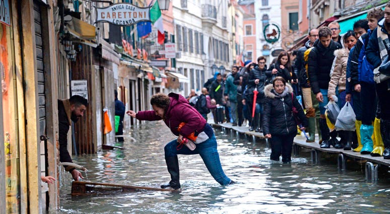 A woman tries to cross a flooded street as people walk on a trestle bridge during high water, in Venice, northern Italy, Friday, Nov. 15, 2019. A woman tries to cross a flooded street as people walk on a trestle bridge during high water, in Venice, northern Italy, Friday, Nov. 15, 2019.