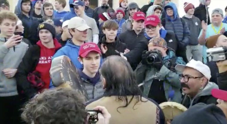 A teenager wearing a "Make America Great Again" hat, center left, stands in front of an elderly Native American singing and playing a drum in Washington. A teenager wearing a "Make America Great Again" hat, center left, stands in front of an elderly Native American singing and playing a drum in Washington.