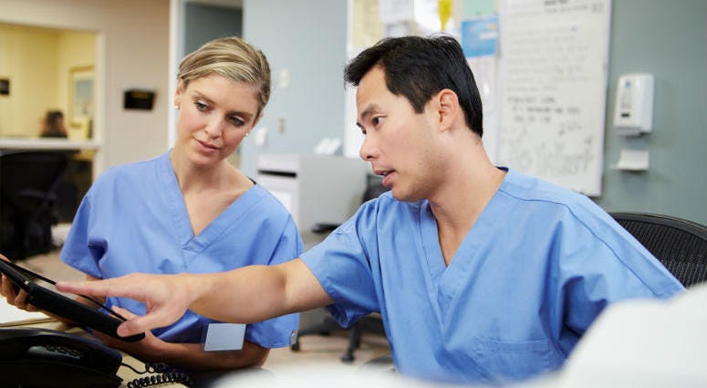 Male And Female Nurse Working At Nurses Station Using Digital Tablet Nurses