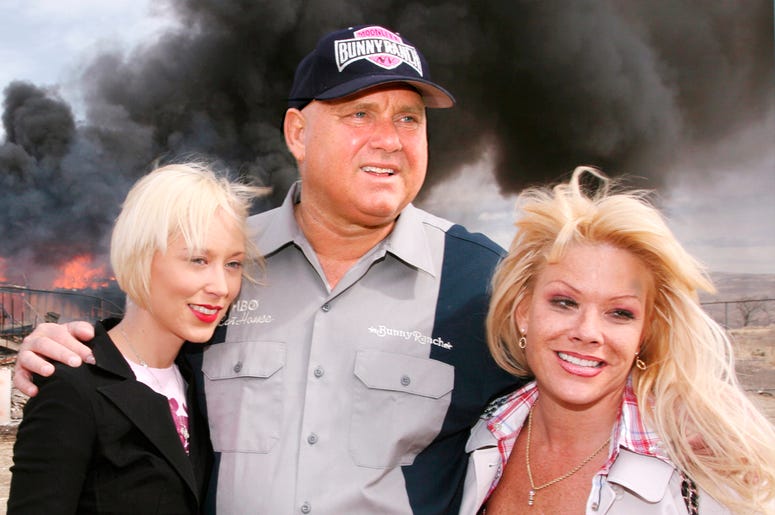 AP18289693349145 In this March 25, 2007, file photo, Moonlite Bunnyranch brothel owner Dennis Hof poses with two of his "working girls" Brooke Taylor, left, and a woman working under the name "Airforce Amy", right, as firefighters burn down remains of the former Mustang R
