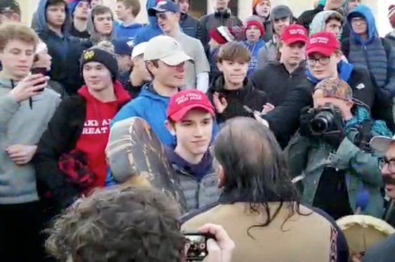 AP19019762910578 In this Friday, Jan. 18, 2019 image made from video provided by the Survival Media Agency, a teenager wearing a "Make America Great Again" hat, center left, stands in front of an elderly Native American singing and playing a drum in Washington. The Roman