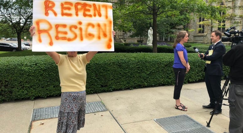 A protester holds a sign outside of St. Joseph's Cathedral in Buffalo after Bishop Malone's press conference. In the background, Siobhan O'Connor speaks with WKBW-TV's Charlie Specht. September 4, 2019 (WBEN Photo/Mike Baggerman)