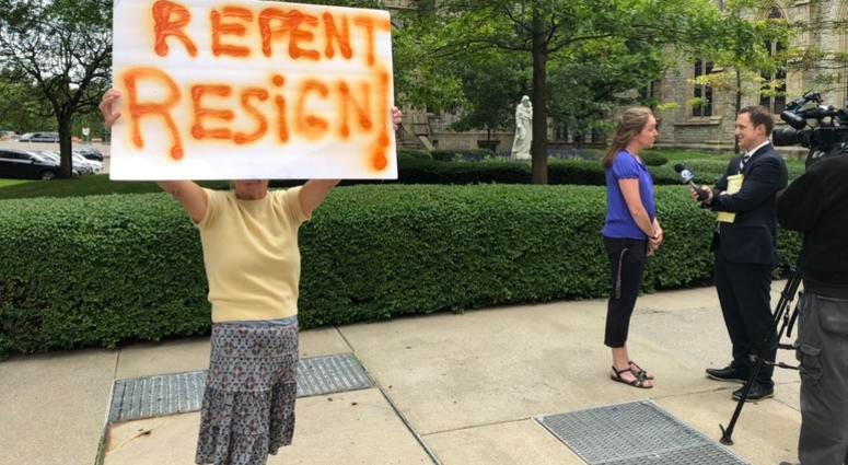 A protester holds a sign outside of St. Joseph's Cathedral in Buffalo after Bishop Malone's press conference. In the background, Siobhan O'Connor speaks with WKBW-TV's Charlie Specht. September 4, 2019 (WBEN Photo/Mike Baggerman)