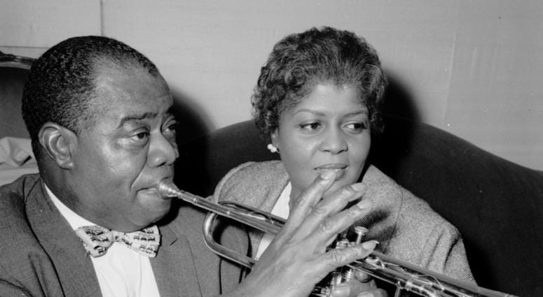 American jazz trumpeter and singer Louis Armstrong (1898 - 1971) plays his trumpet whilst sitting with his wife Lucille in the Mayfair Hotel, London. Louis Armstrong