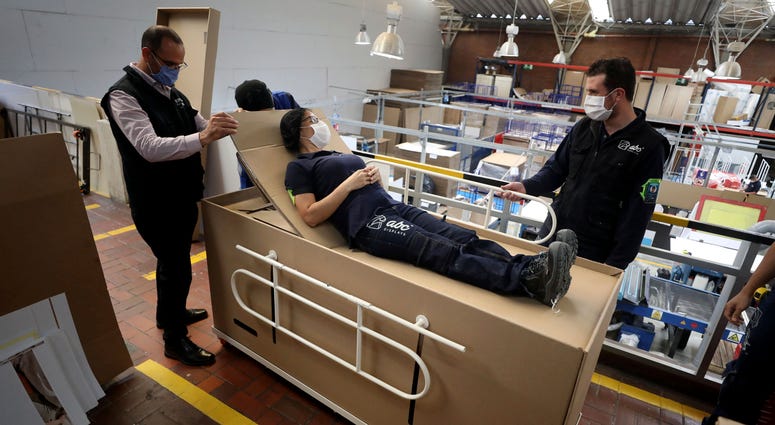 Rodolfo Gomez, to center, and his employees demonstrate how their design of a cardboard box can serve as both a hospital bed and a coffin, designed for COVID-19 patients, in Bogota, Colombia, Friday, May 8, 2020. Coffin hospital beds