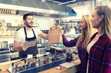 Waiter serving takeaway food to customers at counter in small family eatery restaurant - trendy fast food smiling owner delivering an online to go order in recycled paper bag to clients - stock photo